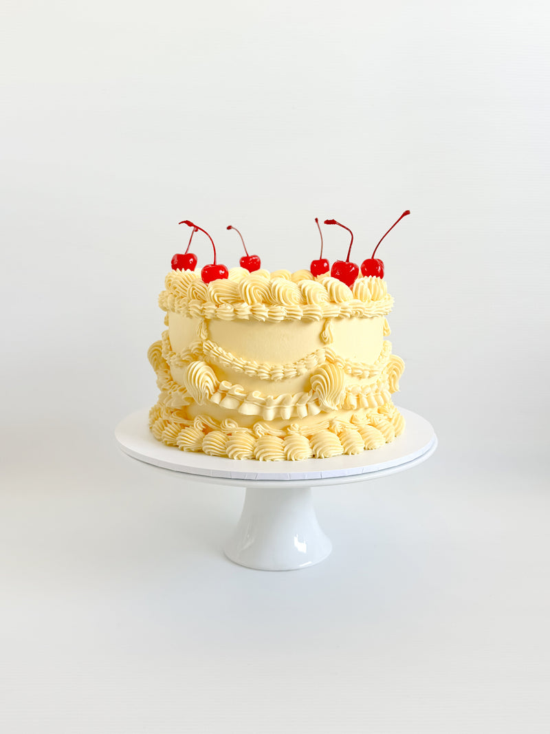 Two-tiered cake with yellow frosting and red cherries on a white stand against a white background