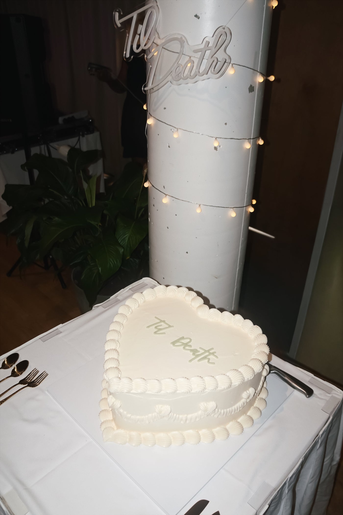 Heart-shaped cake with 'Til Death' text on a white tablecloth, with a tall cylindrical object in the background.